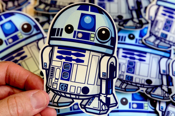 Sticker of R2-D2 from Star Wars held by a hand with multiple R2-D2 stickers in the background.